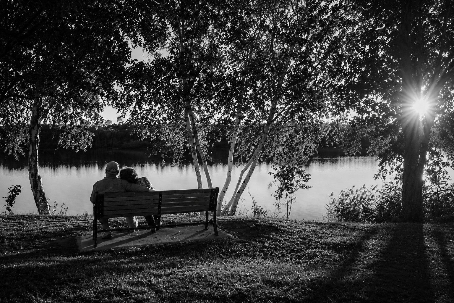 Ett äldre par sitter tätt tillsammans på en parkbänk vid en stilla sjö i kvällsljus — ett svartvitt ögonblick av närvaro, eftertanke och mänskliga möten